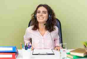 Foto grátis operadora de central de atendimento feminina muito caucasiana sorridente com fones de ouvido, sentada na mesa com ferramentas de escritório isoladas na parede verde