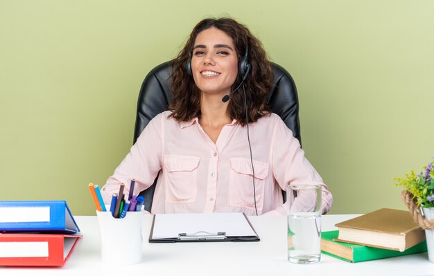 Operadora de central de atendimento feminina muito caucasiana sorridente com fones de ouvido, sentada na mesa com ferramentas de escritório isoladas na parede verde