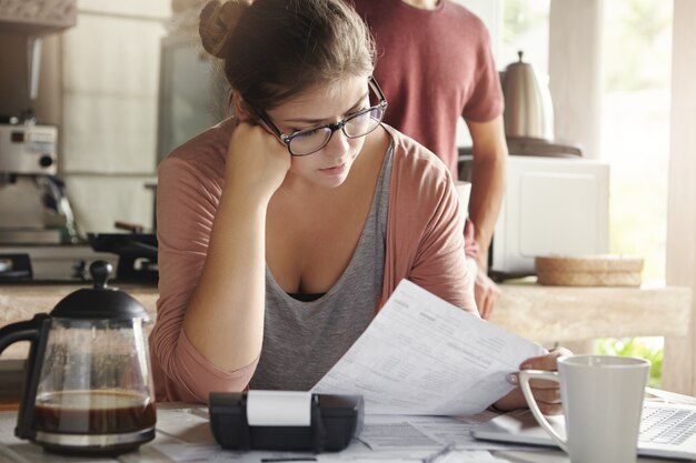 Orçamento e finanças da família. Mulher séria fazendo contas e sentindo-se frustrado com a quantidade de despesas mensais. Jovem fêmea de óculos, cálculo de contas de serviços públicos, sentado à mesa da cozinha