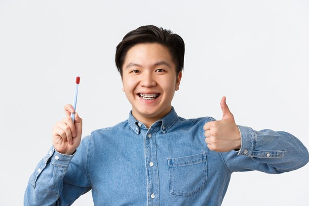 Ortodontia, atendimento odontológico e conceito de higiene. Close de um homem asiático satisfeito mostrando o polegar para cima enquanto recomenda o uso de escova ou pasta de dentes para dentes com aparelho, sorrindo satisfeito