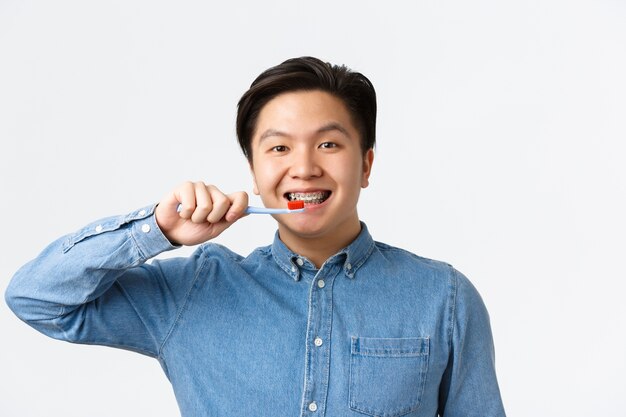 Ortodontia, atendimento odontológico e conceito de higiene. Close de um homem asiático sorridente, de aparência amigável, escovando os dentes com aparelho, segurando uma escova de dentes, em pé na parede branca