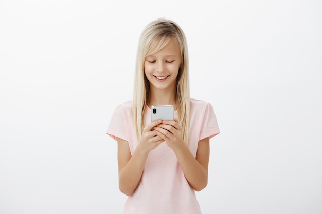 Os amigos ficarão com ciúmes quando virem meu novo telefone. Garoto adorável e alegre e satisfeito com cabelo loiro em uma camiseta rosa, segurando um smartphone, rindo, olhando para a tela, assistindo animação engraçada sobre a parede cinza