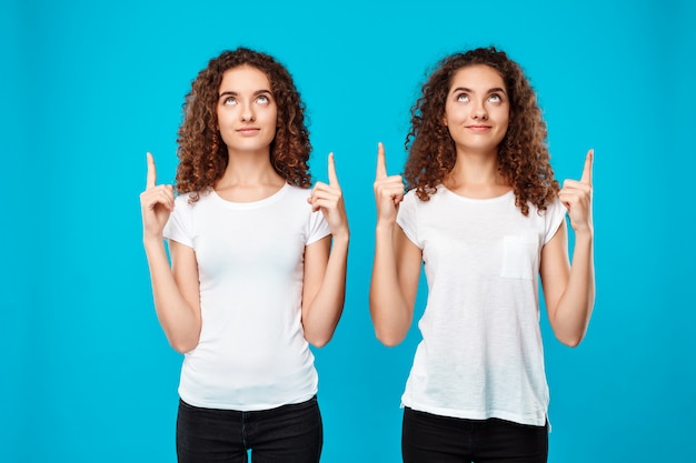 Os gêmeos de duas mulheres que sorriem, apontando os dedos acima sobre o azul.