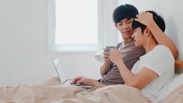 Os homossexuais asiáticos acoplam-se usando o portátil do computador e bebendo o café na casa moderna. Jovem amante da Ásia masculino feliz relaxar descansar juntos depois de acordar, assistindo filme deitado na cama no quarto em casa de manhã