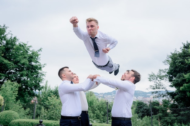 Os melhores homens vestidos em trajes formais estão jogando o noivo como um super-homem, ao ar livre, trabalho em equipe