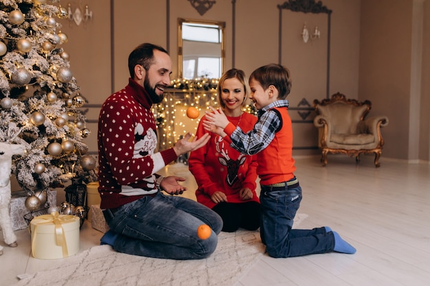 Os pais e seu filho pequeno na camisola vermelha se divertir com laranjas sentado diante de uma árvore de Natal