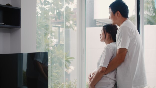 Os pares grávidos asiáticos novos abraçam e guardando a barriga que fala com sua criança. Mamãe e papai se sentindo feliz sorrindo pacífico enquanto cuidar bebê, gravidez perto de janela na sala de estar em casa.