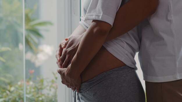 Os pares grávidos asiáticos novos abraçam e guardando a barriga que fala com sua criança. Mamãe e papai se sentindo feliz sorrindo pacífico enquanto cuidar bebê, gravidez perto de janela na sala de estar em casa.