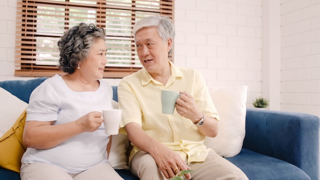 Os pares idosos asiáticos que bebem o café morno e que falam junto na sala de visitas em casa, pares apreciam o momento do amor ao encontrar-se no sofá quando relaxado em casa.