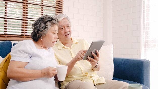 Os pares idosos asiáticos que usam a tabuleta e bebendo o café na sala de visitas em casa, pares apreciam o momento do amor ao encontrar-se no sofá quando relaxado em casa.