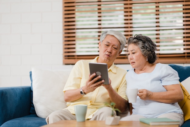 Os pares idosos asiáticos que usam a tabuleta e bebendo o café na sala de visitas em casa, pares apreciam o momento do amor ao encontrar-se no sofá quando relaxado em casa.