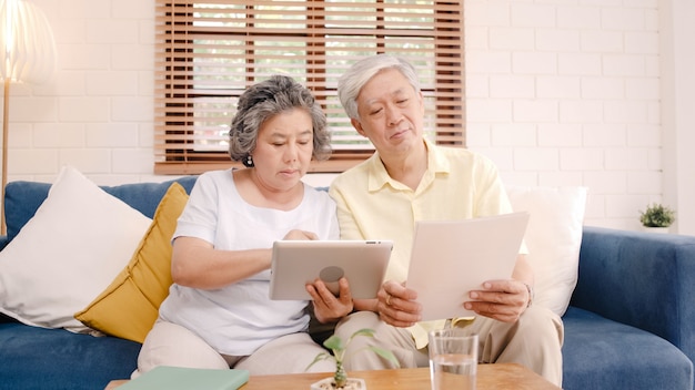 Os pares idosos asiáticos que usam a tabuleta que olha a tevê na sala de visitas em casa, pares apreciam o momento do amor ao encontrar-se no sofá quando relaxado em casa.