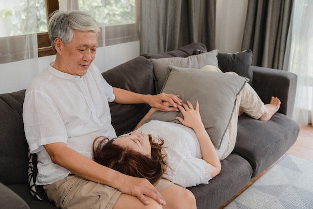 Os pares sênior asiáticos relaxam em casa. As avós chinesas sênior asiáticas, abraço feliz do sorriso do marido encontram-se para baixo seu regaço da esposa ao encontrar-se no sofá no conceito da sala de visitas em casa.