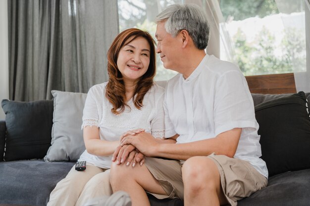 Os pares sênior asiáticos relaxam em casa. O sorriso feliz chinês sênior asiático das avós, do marido e da esposa abraça a fala junto ao encontrar-se no sofá no conceito da sala de visitas em casa.