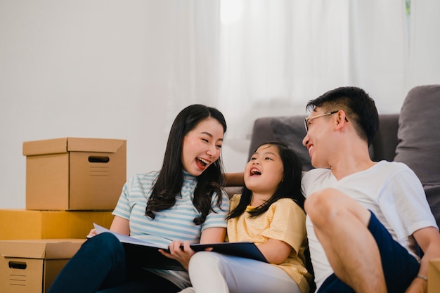 Os proprietários de família novos asiáticos felizes compraram a casa nova. Mãe chinesa, pai e filha abraçando ansiosos pelo futuro em nova casa depois de mudarem-se para a internação, sentada no chão com as caixas juntas.