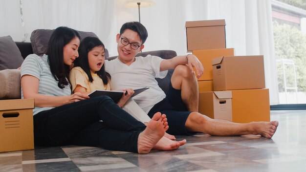 Os proprietários de família novos asiáticos felizes compraram a casa nova. Mãe chinesa, pai e filha abraçando ansiosos pelo futuro em nova casa depois de mudarem-se para a internação, sentada no chão com as caixas juntas.
