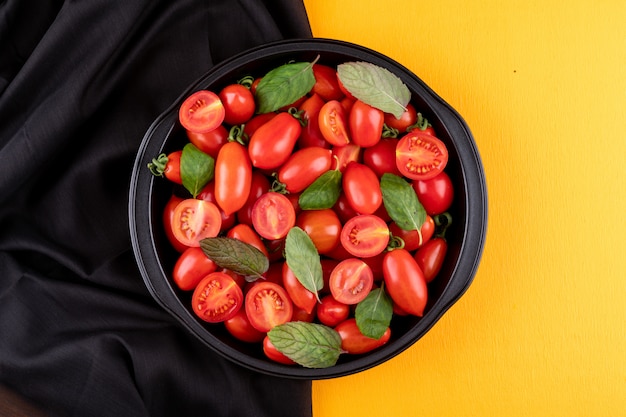 Os tomates-cereja brilhantes vermelhos frescos em panela de metal no pano preto sobre uma superfície amarela