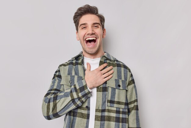 Otimista feliz homem europeu bonito mantém a mão no peito ri com alegria ouve história engraçada vestida de camisa quadriculada expressa emoções positivas isoladas sobre fundo cinza. Conceito de felicidade