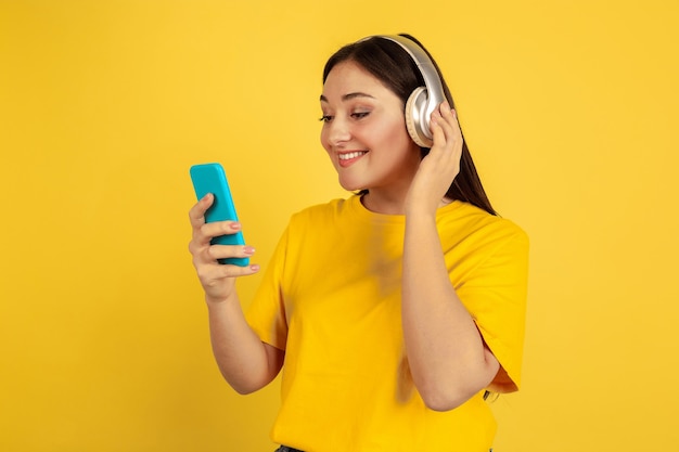 Ouça música com fones de ouvido e telefone sem fio. Mulher caucasiana na parede amarela. Bela modelo Moreno em casual. Conceito de emoções humanas, expressão facial, vendas, anúncio, copyspace.