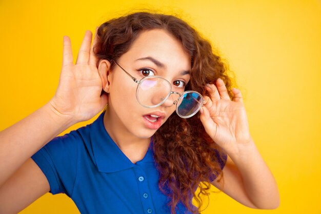 Ouça o segredo. Retrato de menina adolescente caucasiano na parede amarela. Linda modelo feminino encaracolado na camisa. Conceito de emoções humanas, expressão facial, vendas, anúncio, educação. Copyspace.