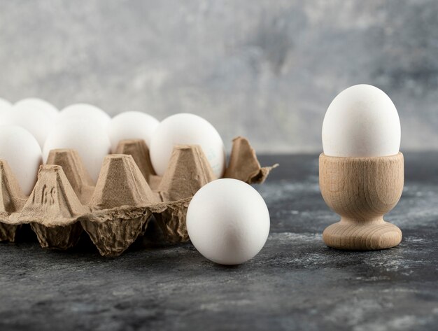 Ovo de galinha cru na taça para ovo com a caixa de ovo numa superfície de mármore.