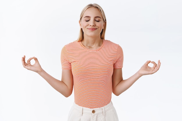 Paciência é a chave para o sucesso. mulher loira atraente, calma e aliviada, caucasiana, com camiseta listrada, olhos fechados e sorridentes reúnem relaxamento, mãos abertas para os lados, faça sinal de mudra, medite, pratique ioga