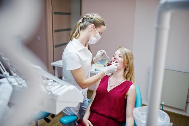 Paciente atraente em vestido vermelho violeta deitado na cadeira odontológica enquanto dentista feminina tratando seus dentes com instrumentos especiais