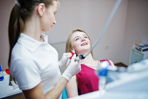 Paciente atraente em vestido vermelho violeta deitado na cadeira odontológica enquanto dentista feminina tratando seus dentes com instrumentos especiais