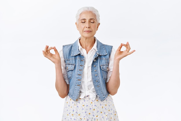 Foto grátis paciente, calma e saudável avó moderna com cabelos grisalhos penteados em um elegante colete jeans, vestido, meditando em pé na ioga, pose de mudra, olhos fechados, respiração de alívio de estresse