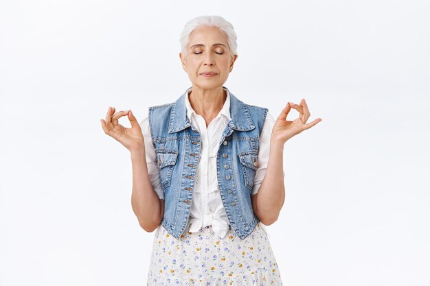 Paciente, calma e saudável avó moderna com cabelos grisalhos penteados em um elegante colete jeans, vestido, meditando em pé na ioga, pose de mudra, olhos fechados, respiração de alívio de estresse