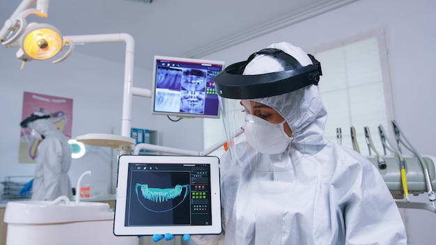 Paciente com explicação de escuta para tratamento dentário, dentista de macacão mostrando raio-x no tablet. Especialista em estomatologia usando traje de proteção contra infecção por covid19 apontando para a radiografia