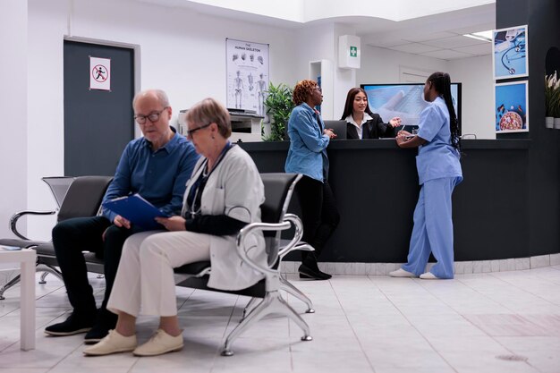 Paciente e enfermeira sentados na recepção, conversando com recepcionista feminina sobre diagnóstico de doenças e suporte de saúde. Diversas pessoas trabalhando no centro de saúde no balcão de registro.