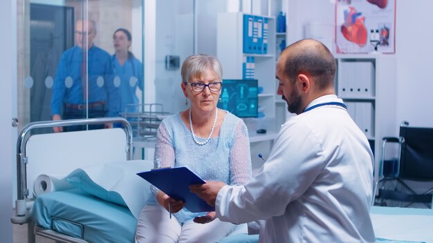 Paciente sênior, assinando o formulário de decisões médicas, sentado na cama do hospital em uma clínica privada moderna. Médico com área de transferência, enfermeira trabalhando em backgorund. Documentos do sistema médico-medicinal de saúde contra