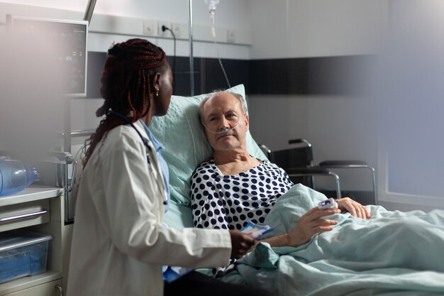 Paciente sênior doente, deitada na cama, respirando por meio de tubo de ensaio de oxigênio, ouvindo o médico