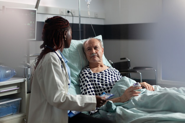 Paciente sênior doente, deitada na cama, respirando por meio de tubo de ensaio de oxigênio, ouvindo o médico