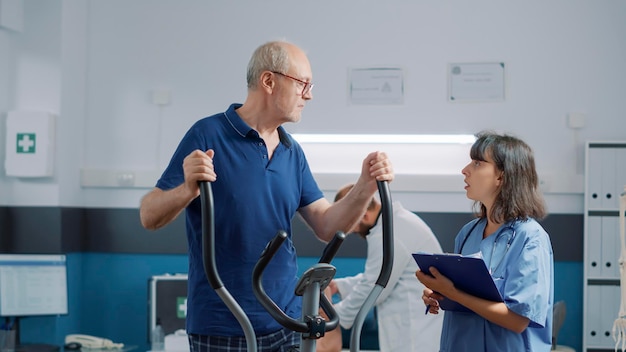 Paciente sênior fazendo exercício de recuperação no gabinete médico, recebendo assistência de especialista em reabilitação. Enfermeira explicando o uso de bicicleta estacionária para recuperação de fisioterapia.