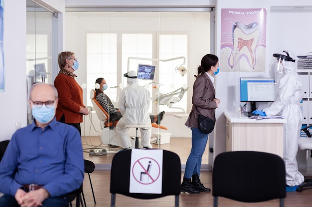 Pacientes com máscara de proteção aguardando na recepção de estomatologia com novo normal, equipe vestindo traje ppe