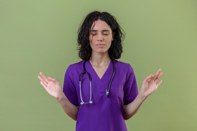 Pacífica jovem enfermeira vestindo uniforme e estetoscópio praticando exercícios respiratórios de ioga com os olhos fechados, em pé no verde isolado