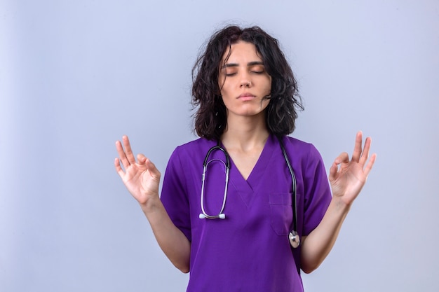 Pacífica jovem enfermeira vestindo uniforme e estetoscópio praticando exercícios respiratórios de ioga com os olhos fechados em pé