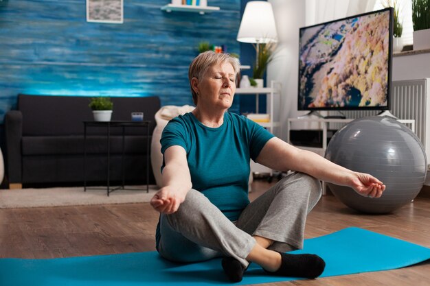 Pacífica mulher sênior com os olhos fechados, sentada na esteira de ioga, meditando durante o treino de bem-estar. Pensionista confortável praticando posição de lótus e exercitando concentração corporal na sala de estar