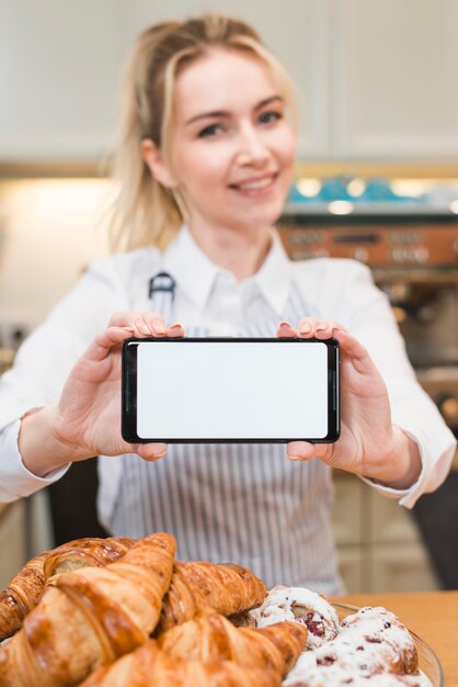 Padeiro feminino mostrando telefone inteligente com tela branca em branco perto do croissant assado