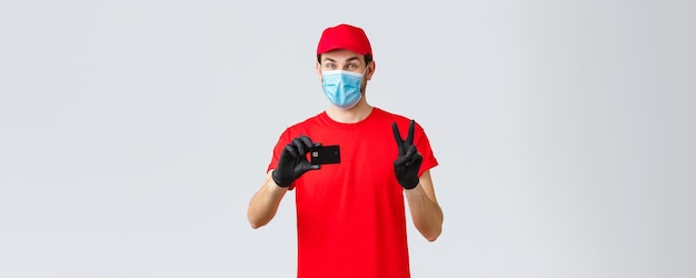 Foto grátis pagamento de entrega sem contato e compras on-line durante a quarentena covid19 correio amigável em luvas de uniforme vermelho e máscara facial fornecem opção de pagamento segura mostrar cartão de crédito e sinal de paz