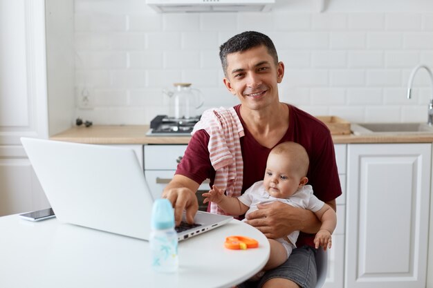 Pai adulto jovem sorridente feliz vestindo camiseta casual marrom, sentado à mesa na cozinha perto do notebook, segurando o bebê nos braços, olhando para a câmera com expressão positiva.