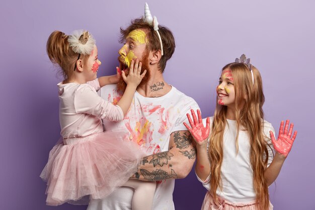 Pai afetuoso e chocado está com o rosto sujo de tintas, abre a boca amplamente, segura a filha nas mãos. menina sorridente usa coroa, mostra as palmas das mãos em aquarelas cor de rosa. pai alegre e filhos se divertem