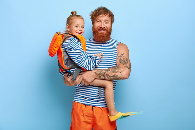 Foto grátis pai afetuoso feliz carrega a filha pequena nas mãos que usa colete salva-vidas protetor e nadadeiras, indo nadar juntos, aproveitar o verão, ter cabelo ruivo. família ruiva de férias