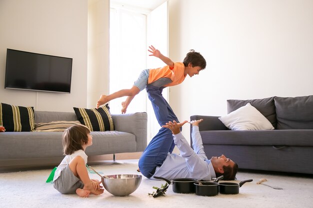 Pai alegre segurando o filho nas pernas e deitado no tapete. Menino caucasiano feliz voando na sala de estar com a ajuda do pai. Garoto bonito sentado no chão perto de uma tigela e panelas. Conceito de infância e fim de semana