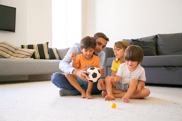 Pai concentrado segurando uma bola e conversando com as crianças. Amoroso pai caucasiano e filhos sentados no tapete da sala e brincando juntos. Infância, atividade lúdica e conceito de paternidade