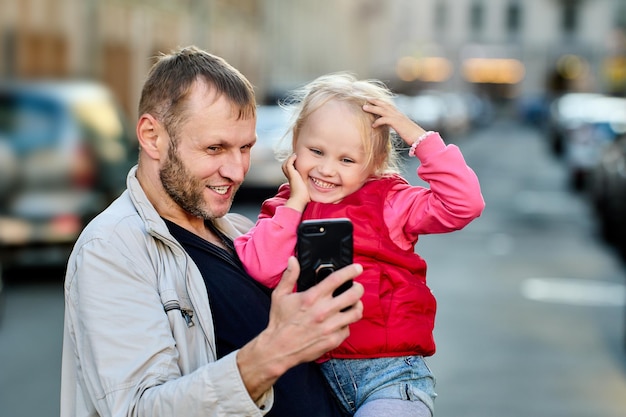 Pai e filha com a mão estão assistindo a vídeos e sorrindo na rua com trânsito.