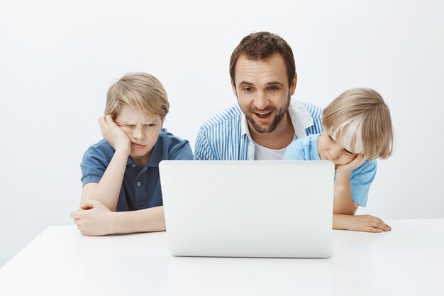 Pai engraçado satisfeito sentado com os filhos perto do laptop, olhando para a tela com um sorriso feliz e satisfeito, enquanto os meninos se sentem entediados e indiferentes