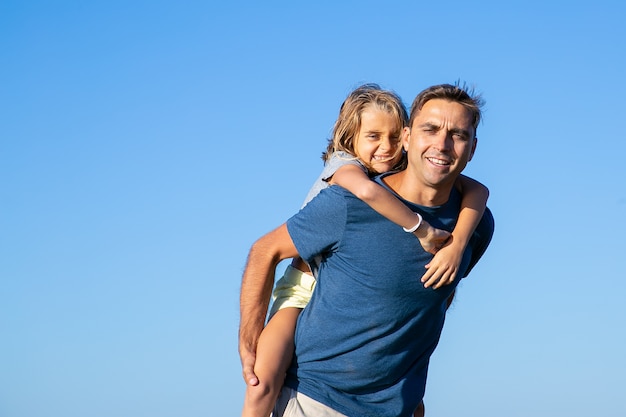 Foto grátis pai feliz carregando uma garota alegre nas costas. pai e filha, aproveitando o tempo de lazer juntos ao ar livre. família e conceito de caminhada ao ar livre